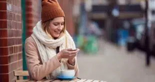 Female Customer Sending Text Message Sitting Outside Coffee Shop On Busy City High Street In Fall
