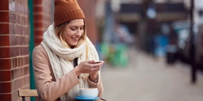 Female Customer Sending Text Message Sitting Outside Coffee Shop On Busy City High Street In Fall