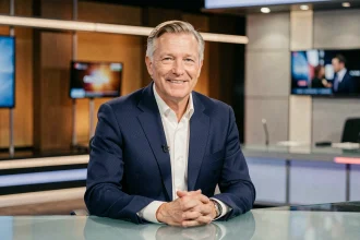 Présentateur TV français emblématique assis derrière un bureau de plateau de télévision, en costume bleu marine, regard confiant face caméra
